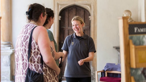 Three people are standing in a group. One is smiling at the other two and has her arm out in a welcoming gesture.